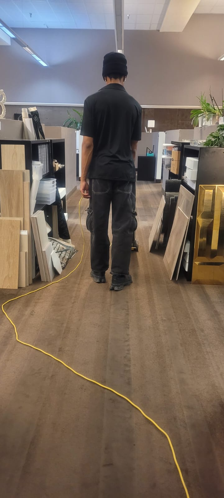 Person wearing black clothing standing in a room with wood and tile samples on shelves, holding a cord attached to a floor polishing machine.