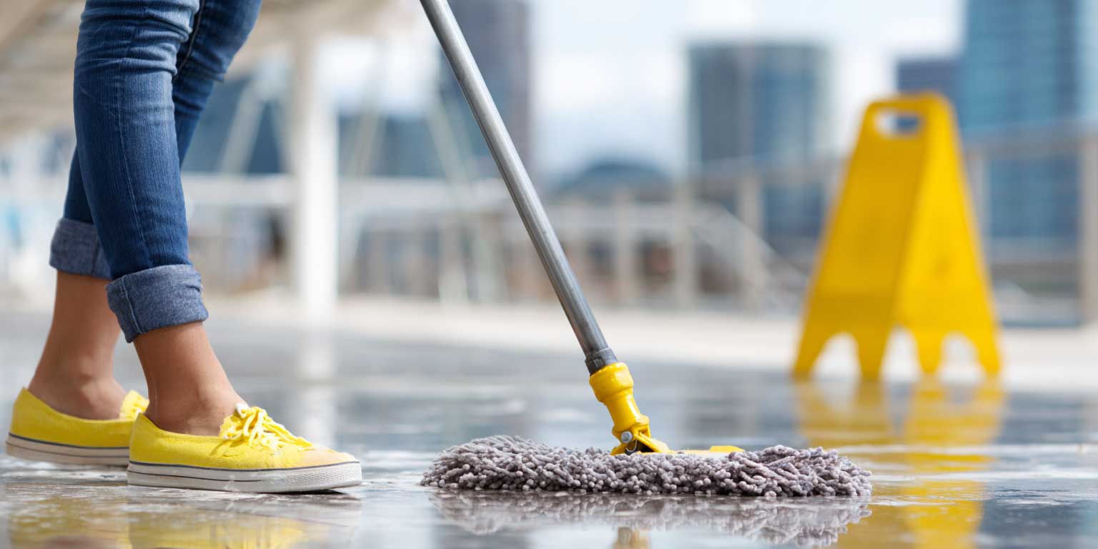 Yellow push broom sweeping a shiny wet tiled floor in a spacious room with large windows.